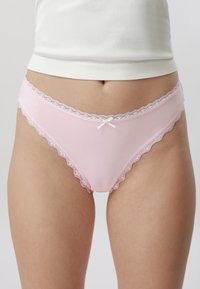 Žena v béžovo růžovém spodním prádle s krajkovou vsadkou ve stylu bikin a bílém topu, zobrazena od pasu po horní část stehen.