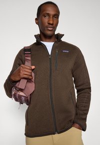 Brown zip-up fleece jacket with side pockets, high collar, and black accents. Individual holds a small, pink shoulder bag.
