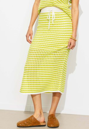 Femme portant une jupe tricotée jaune et blanche à motif avec une taille à cordon et des chaussures marron à enfiler, debout contre un mur blanc.