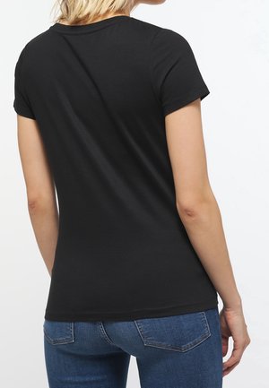Žena v jednoduchém černém tričku s krátkým rukávem a modrých džínách, zobrazena zezadu na bílém pozadí.