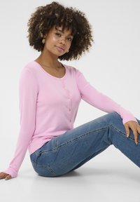 Lichtroze lange mouwen cardigan met een ronde hals en vijf gouden knopen, gedragen met een paar blauwe denim jeans, zittend tegen een neutrale achtergrond.