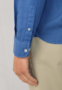 Blauwe katoenen shirt met een keurig kraag, knoopsluiting aan de mouwen en een gladde textuur; gecombineerd met lichtbeige broek, met een hand die op het been rust.