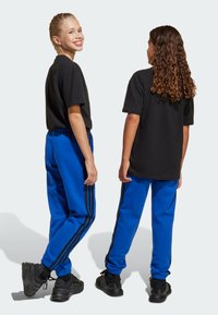 T-shirts noirs à manches courtes associés à des survêtements bleus avec des rayures noires sur les côtés. Les deux portent des chaussures de sport noires.