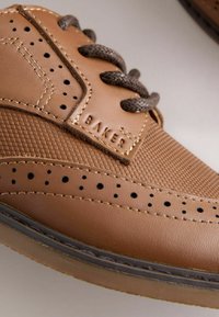 Zapato de cuero marrón con superficie texturizada y detalles perforados. Cuenta con cordones marrón oscuro y una etiqueta "BAKER". Suela de goma con borde en contraste.