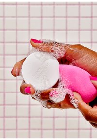 Biała myjąca kostka mydła oraz różowa gąbka do makijażu są trzymane w dłoniach, otoczone bąbelkami mydła, na różowym tła w kratkę.