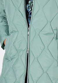 Fodrad pufferjacka i ljusgrön, med dragkedjestängning och diamantmönster. Materialet är slätt med en lätt vikt.
