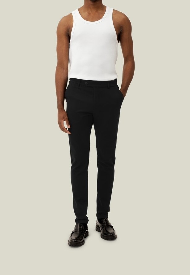 Homme portant un débardeur blanc sans manches, un pantalon noir slim et des mocassins noirs, debout avec une main dans la poche.