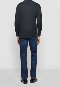 Musta pitkähihainen paita korkealla kauluksella, yhdistetty tummansinisiin farkkuihin. Kengät ovat kiiltävää mustaa nahkaa. Kuvattu takaa.