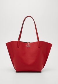 Červená taška přes rameno s hladkým, lesklým povrchem. Obsahuje dvě pevné ucha a stříbrnou kovovou sponu. Logo "GUESS" prominentně zobrazeno.