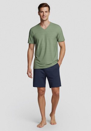 Uomo in piedi a piedi nudi che indossa una maglietta verde a maniche corte con scollo a V e pantaloncini blu scuro su uno sfondo chiaro uniforme.