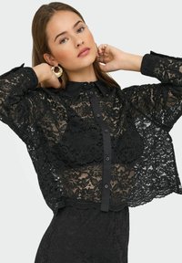 Blouse en dentelle noire avec un devant à boutons, un col pointu et des manches transparentes. Présente des motifs floraux et un design texturé sur l'ensemble.