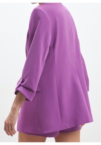 Lila blazer med lös passform, featuring uppvikta ärmar, en mjuk textur och en enda knappdetalj på ärmsluten.