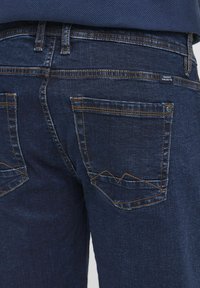 Jean en denim bleu foncé avec deux poches arrière à coutures angulaires, couture visible en orange et une petite étiquette de marque sur la ceinture.