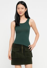 Jauna sieviete ar garām melnām matiem, ģērbusies tumši zaļā topā un tumši zaļā kantainā svārkos ar lieliem priekšējiem kabatām.