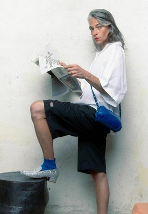 Pantaloni corti neri fino al ginocchio, camicia bianca oversize, borsa a tracolla blu, calzini turchesi e scarpe glitterate argento. Tiene in mano una rivista.