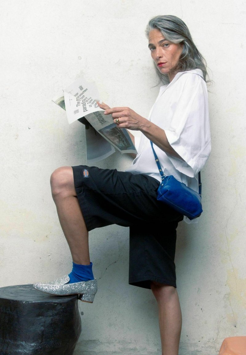 Černé kraťasy ke kolenům, oversized bílá košile, modrá crossbody taška, tyrkysové ponožky a stříbrné třpytivé boty. Drží časopis.