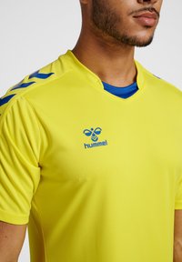 Gul sportströja med blå accenter och logotyp, korta ärmar, V-ringning, slät textur, med kontrasterande blå krage och axeldetaljer.