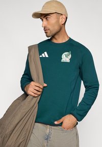 Μπλουζάκι με μακριά μανίκια σε τυρκουάζ χρώμα με το έμβλημα του Μεξικού και λευκές ρίγες Adidas, συνδυασμένο με ένα ανοιχτό καφέ γιλέκο. Το μοντέλο κρατά το γιλέκο άνετα.