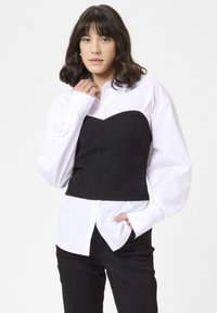 Sort struktureret korset top lagdelt over en hvid skjorte med knapper, der har lange ærmer og en tætsiddende design, hvilket skaber en moderne kontrast.