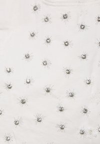 Vit tyg dekorerad med silverbroderi i en solstrålemönster, med små runda prydnader i mitten av varje stråle.