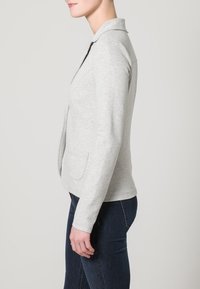 Szary, miękki materiałowy blezer z kołnierzem, bocznymi kieszeniami i dopasowanym krojem, pokazany z boku na jednolitym tle.