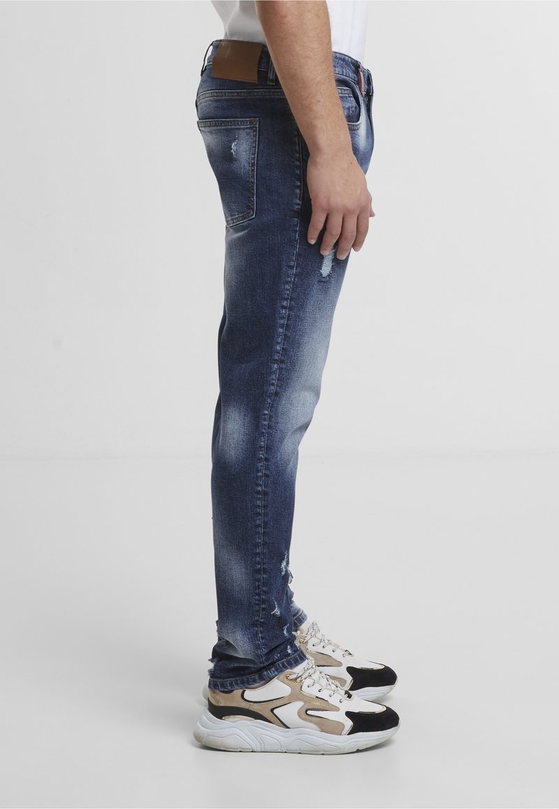 Homme portant un jean bleu délavé et usé ainsi que des baskets blanches, beiges et noires, debout de profil.