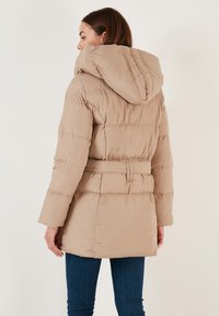 Béžová péřová bunda s kapucí, opásaným pasem a prošívanými panely. Obsahuje dlouhé rukávy a design do poloviny stehen.