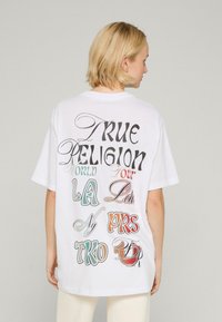 Biała, oversize'owa koszulka z krótkim rękawem, z klasycznym okrągłym dekoltem, z kolorowym graficznym napisem na plecach: "TRUE RELIGION WORLD TOUR LA NYC PRS TKO."