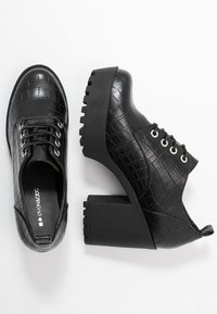 Zapatos de cuero negro con textura de cocodrilo, puntera redonda, diseño de cordones y un tacón macizo. Suela de goma con detalles de relieve.