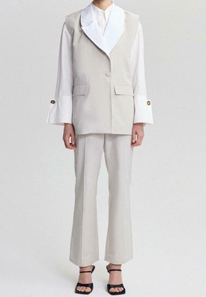 Person wearing beige tailored vest and pants with white long-sleeve shirt featuring wide cuffs with gold buttons, paired with black open-toe heels.