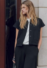 Schwarze kurz geschnittene Jacke mit kurzen Ärmeln, großen vorderen Taschen und Knopfverschluss, getragen über einem weißen Rippoberteil und einer schwarzen Hose.