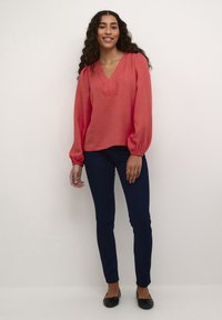 koralová textúrovaná blúzka s výstrihom do V a dlhými puffovými rukávmi, skombinovaná s tmavomodrými slim-fit džínsami a čiernymi plochými topánkami.
