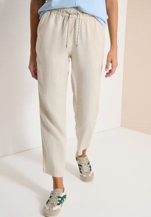 Personne portant un pantalon beige à cordon et des baskets beiges avec des rayures vertes, debout sur un sol blanc devant des murs neutres.