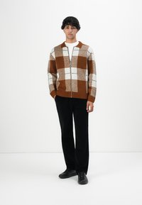 Junger Mann, der einen braun-weißen karierten Zip-Pullover, ein weißes Hemd, schwarze Hosen und schwarze Schuhe trägt, vor einem einfarbigen Hintergrund steht.