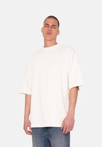 Camiseta blanca de gran tamaño con cuello redondo y mangas cortas, que presenta un sutil logo de la marca en el frente. Llevada con vaqueros azules.