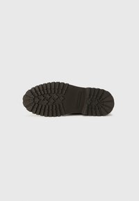 Suela de bota de goma negra con dibujos profundos y una sección central lisa; presenta detalles en patrón cruzado para una mejor adherencia y durabilidad.