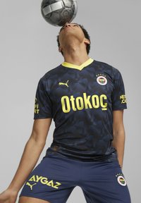 Sotilas sininen jalkapallopaita keltaisilla yksityiskohdilla ja logolla, jossa on geometrinen kuvio. Lyhyet hihat, yhdistettyinä vastaaviin housuihin. Jalkapallo tasapainotettuna päällä.