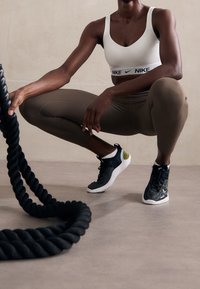 Svart rep på en slät yta, modell i vit sport-bh med Nike-logga, bruna leggings och svart/vita träningsskor med grön accent.