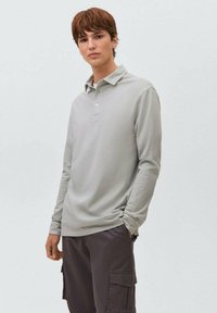 Giovane uomo con capelli corti castani che indossa una polo a maniche lunghe grigio chiaro e pantaloni cargo scuri su uno sfondo neutro.