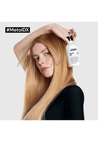 Bouteille de shampooing Metal DX tenue contre des cheveux blonds longs et raides. La bouteille est blanche avec un texte noir. Le modèle porte un haut noir.