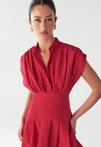 Rotes Kleid mit einem geknöpften Kragen, kurzen Puffärmeln und taillierter Passform. Verfügt über einen plissierten Rock und eine glatte Textur.