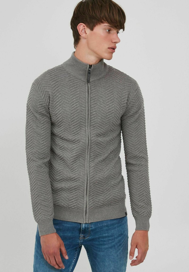 Grå zip-up sweater med et struktureret chevronmønster, ribbet krave og manchetter samt en slim fit. Båret over denim jeans.