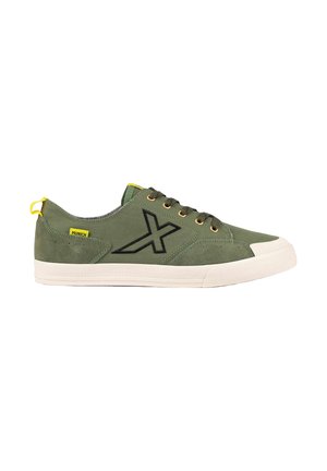 Zapatilla baja de color verde oliva con logo negro en forma de X, suela de goma blanca, lengüeta amarilla y etiqueta amarilla a juego en el lateral.