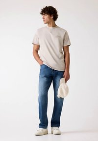T-shirt en coton beige à manches courtes avec un détail brodé, associé à un jean en denim bleu. Baskets blanches et une casquette légère tenue à la main.