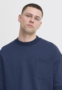 Tumesinine sweatshirt ribitud ümarate kaelustega, lühikeste varrukatega ja rinnataskuga. Valmistatud tekstuurist kangast, sirge lõike ja minimaalset disaini omavad.