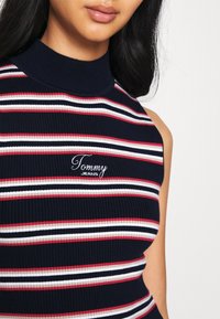 Pruhovaný bezrukávový top s vysokým límcem. Barvy: námořnická modř s červenými a bílými pruhy. Dokončeno vyšívaným logem "Tommy Jeans".