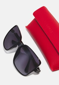 Black oversized sunglasses with dark lenses, accompanied by a textured red leather case embossed with the brand name "GUESS."