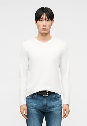 Mlad moški s ravnimi črnimi lasmi, oblečen v belo majico z dolgimi rokavi in V-izrezom ter modre kavbojke s črnim pasom, stoji pred enobarvnim ozadjem.