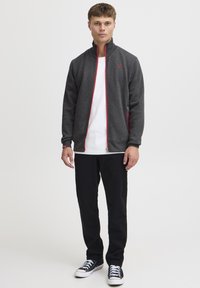 Tumehall lukuga jope punaste detailidega, halli kangaga struktuur, kõrge krae ja rindkere peal olev logo, kantud valge T-särgi ja mustade pükstega.