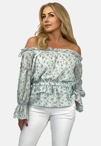 Jasnoniebieska bluzka w kwiaty z odkrytymi ramionami, z falbaniastymi rękawami, elastycznym pasem i falbaniastymi detalami. Połączona z białymi spodniami. Wzór florystyczny na całej powierzchni.